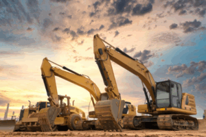 Two men discussing plans at a construction site; one wearing a blue hard hat and casual attire, the other in business clothes holding a green clipboard, with heavy machinery in the background.
