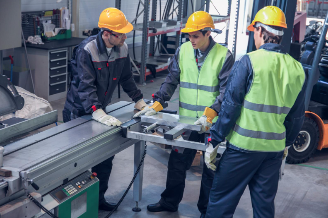 Three factory workers wearing safety helmets, gloves, and reflective vests operating machinery together in an industrial workshop.