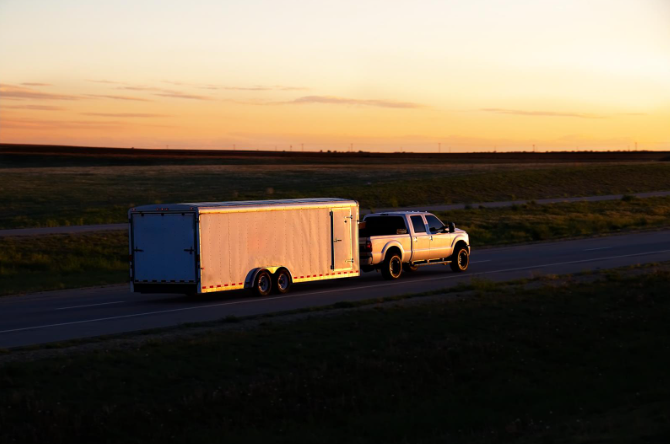 A pickup truck towing a large enclosed trailer along a rural highway at sunset, with warm light reflecting off the trailer's side.