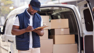 A smiling delivery worker wearing a cap and vest writes on a clipboard while standing beside a van loaded with cardboard boxes.