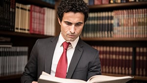 Professional man in a suit with a red tie reading an open book in a law library, surrounded by shelves filled with legal volumes and reference books, conveying focus and professionalism.