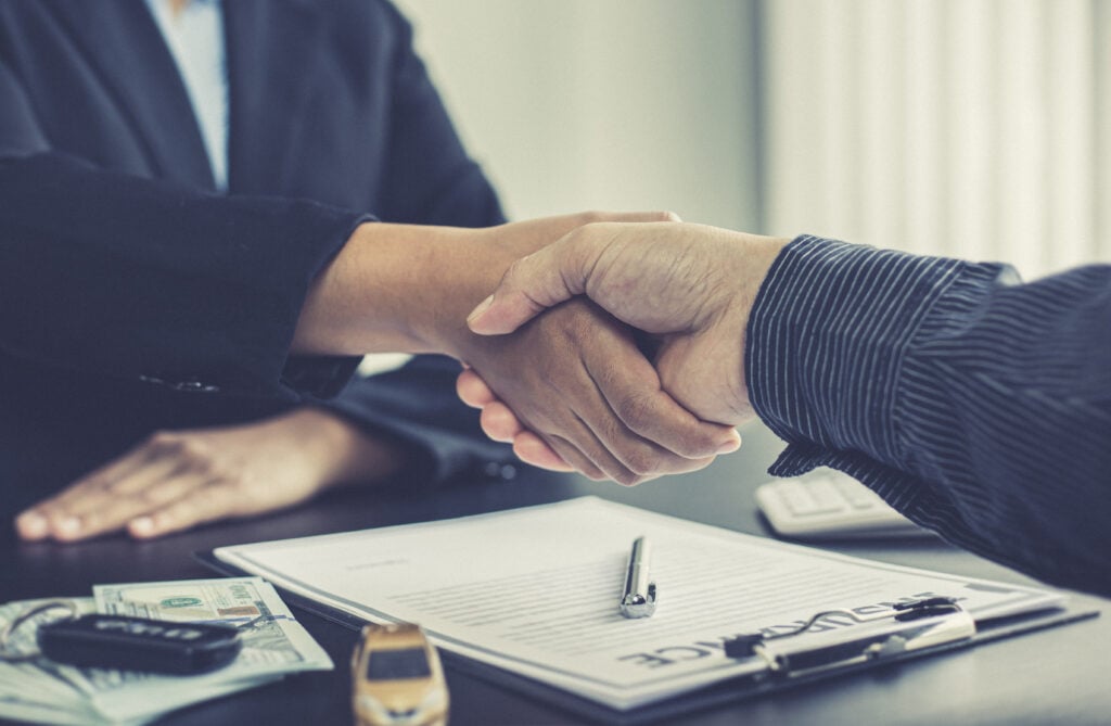 Two people shaking hands over a signed contract on a desk, with a pen, cash, and a car key visible, symbolizing a legal or financial agreement.