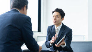 Two business professionals in suits seated in an office, discussing documents as one gestures with a tablet during a formal meeting.