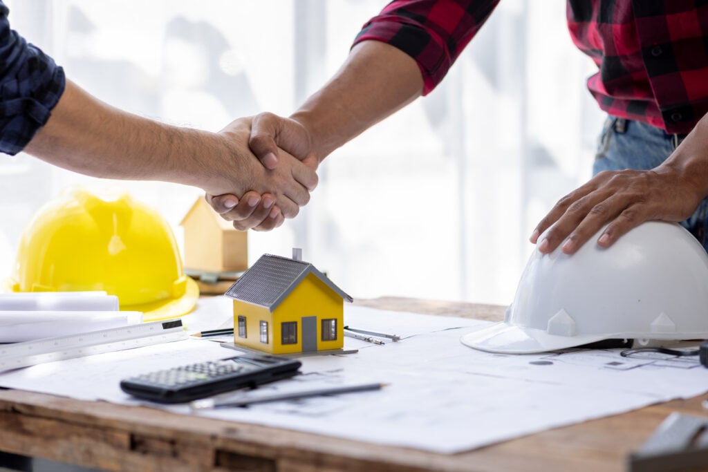 Two construction professionals shake hands over blueprints on a table with hard hats, tools, and a small model house, symbolizing an agreement.