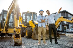 Two construction professionals wearing hard hats standing in front of multiple yellow excavators and loaders at an equipment yard, with one pointing while discussing project details