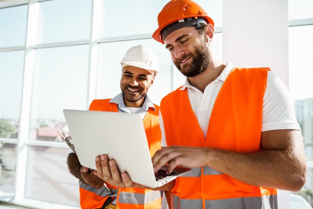 Two male construction workers wearing orange safety vests and hard hats reviewing project plans on a laptop near a bright window, collaborating on site management and documentation