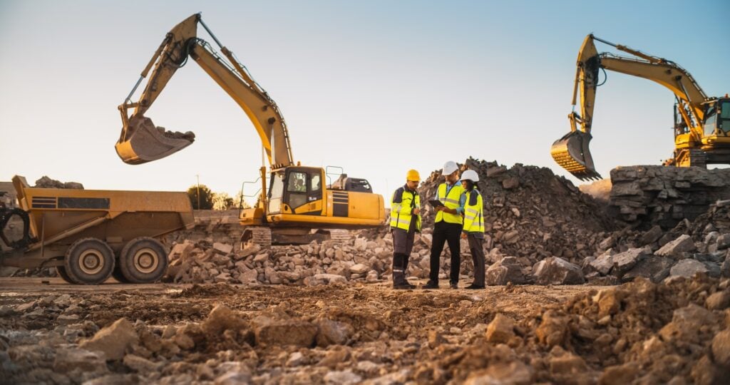 Three construction workers wearing hard hats and orange safety vests standing on a gravel site, surrounded by operating excavators and dump trucks, managing heavy equipment operations at a quarry or mining facility