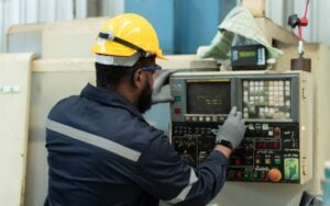 Industrial worker wearing a yellow hard hat, safety glasses, and protective gloves operates a CNC machine control panel in a manufacturing facility.