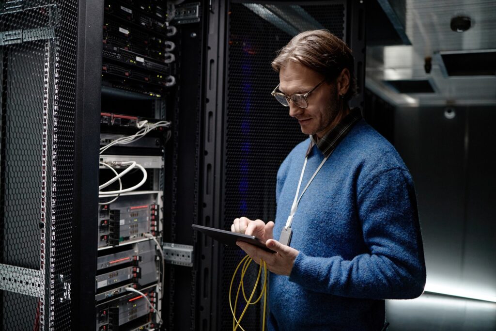 IT professional with glasses and a security badge uses a tablet to monitor server hardware in a data center, with network cables visible in the rack.
