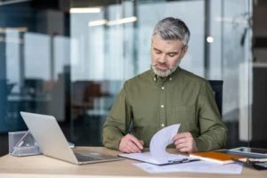 Mature businessman in olive shirt carefully reviewing financing contract documents at an office desk with laptop