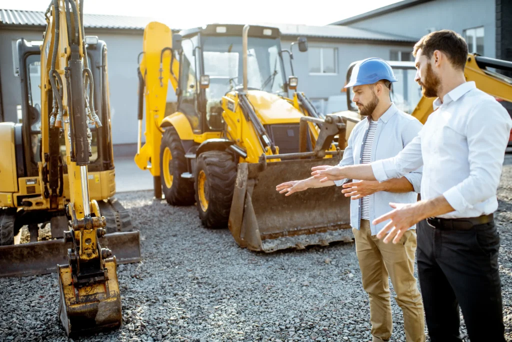 Two professionals in casual workwear discussing equipment options in front of a yellow excavator and backhoe loader