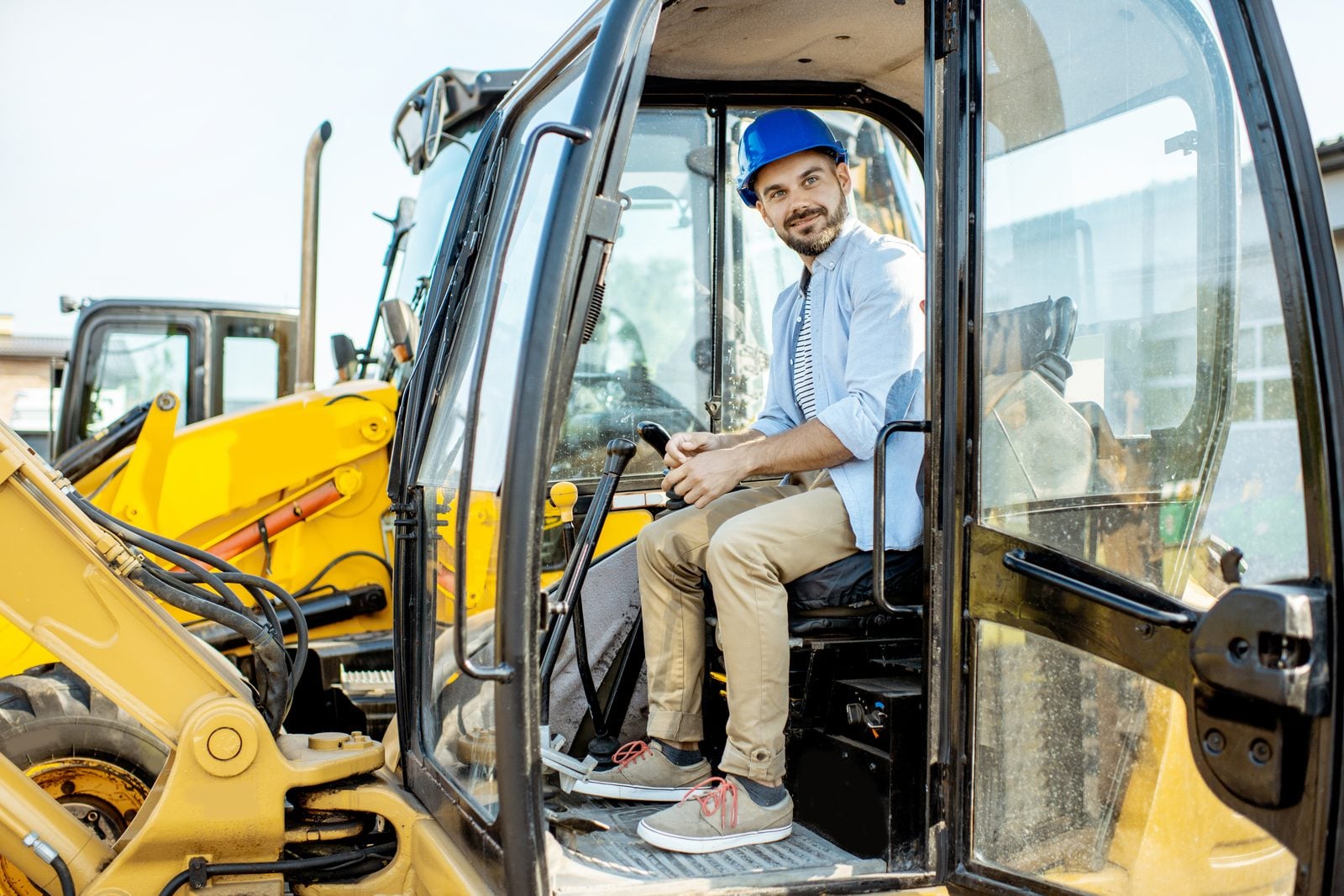 Business owner in a hard hat seated in the cab of a yellow excavator at an equipment yard